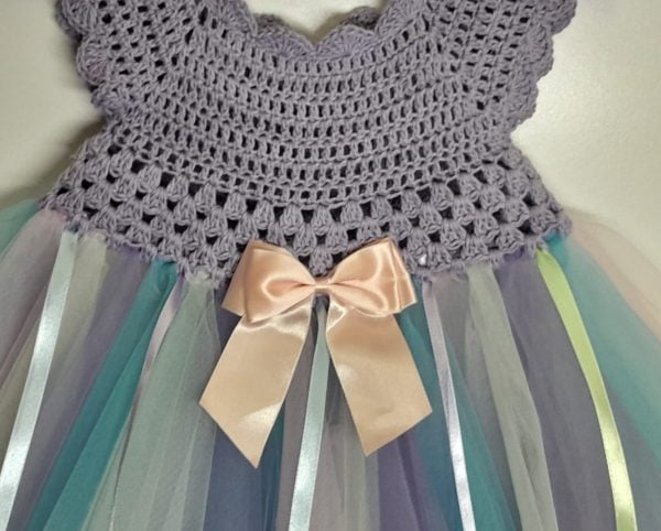 Close-up of a crocheted toddler dress bodice in soft grey-purple cotton, featuring delicate detailing and a pastel pink satin bow at the center.