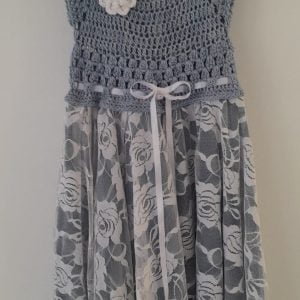 Handcrafted crocheted grey cotton dress with white floral printed tulle skirt This image shows a dress for a toddler aged 3 years. The bodice is crocheted from a soft grey cotton yarn. It has short capped sleeves and is embellished with a single white crocheted flower. A subtle white satin ribbon accentuates the neckline. The full skirt is made of layered wispy grey tulle with an overlay of a white chevron print featuring delicate rose motifs. The blended fabrics and heritage craftsmanship conveys a spirit of gentle nurture and grace. Photographs of a child dressed in this handmade keepsake will preserve precious milestones, discoveries unfolding softly across seasons wrapped in moments treasured forever in living memory.